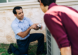 Aire Serv tech kneeling next to air conditioning unit explaining a part to a customer.