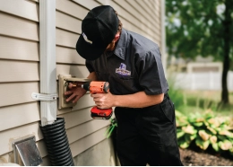A technician is using a power drill to install a fixture on the side of a house.