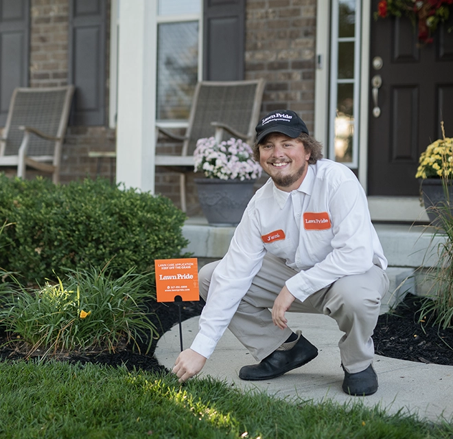 A lawn care worker kneeling on a front walkway next to a lawn service sign outside a house.