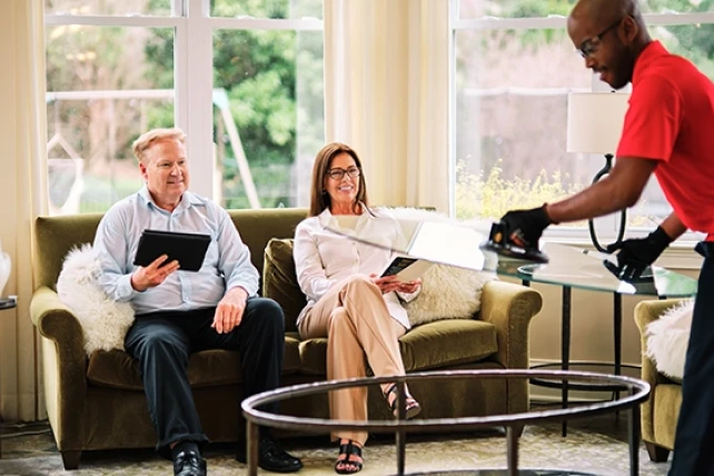 A man and a woman sit on a couch while a worker adjusts a glass table nearby.