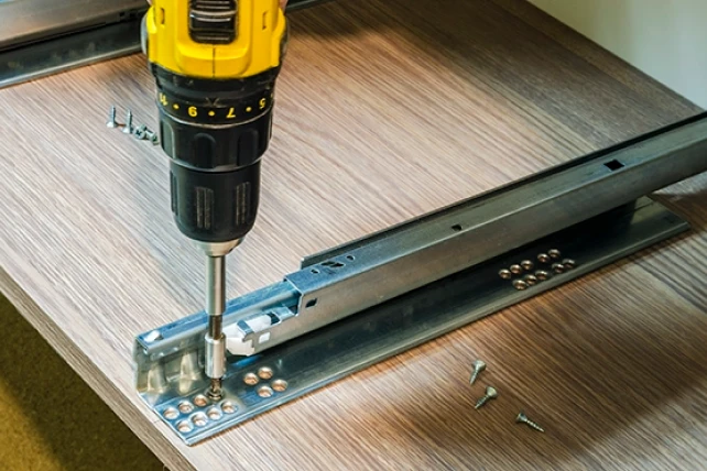 A power drill attaching a metal drawer slide to a wooden panel.