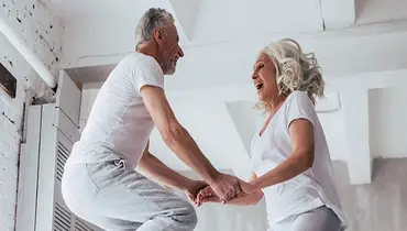 Older couple dressed in white jumping on a bed.