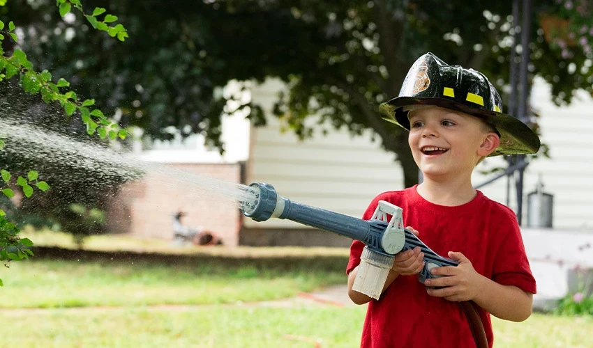 Child spraying water out of a hose and wearing firefighter helmet.