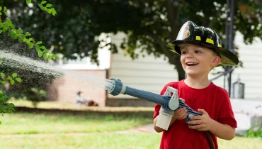 Child spraying water out of a hose and wearing firefighter helmet.