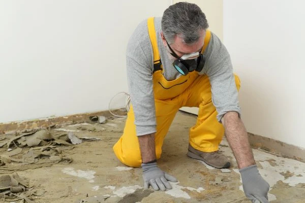 A man in a yellow jumpsuit wearing a respiratory removing carpet adhesive from a floor.