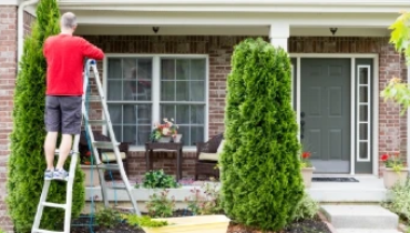 Man in a red shirt on a ladder trimming tree in front yard of a residential home.