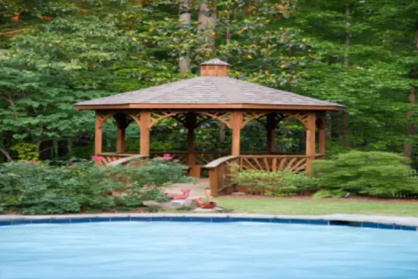 A beautiful inground pool surrounded by greenery and a wooden gazebo.