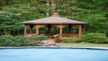 A beautiful inground pool surrounded by greenery and a wooden gazebo.