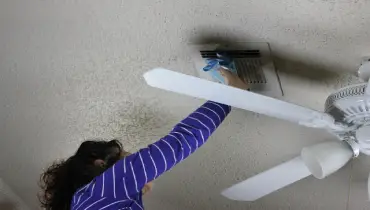 woman cleaning ceiling air ducts near fan.