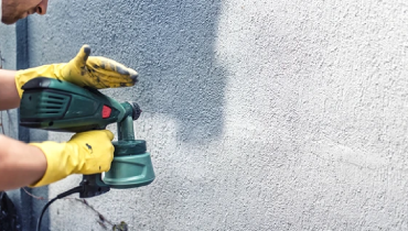 Person using a paint sprayer on the exterior of a home.
