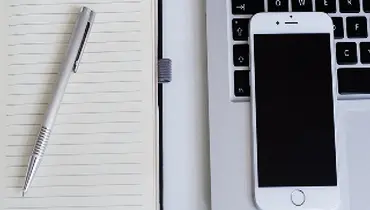 Pen and iPhone on a desk with notepad and laptop computer.
