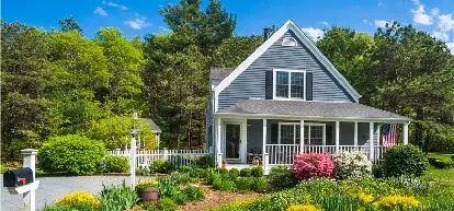 Two story home with front porch and flowers in front and surrounded by trees.